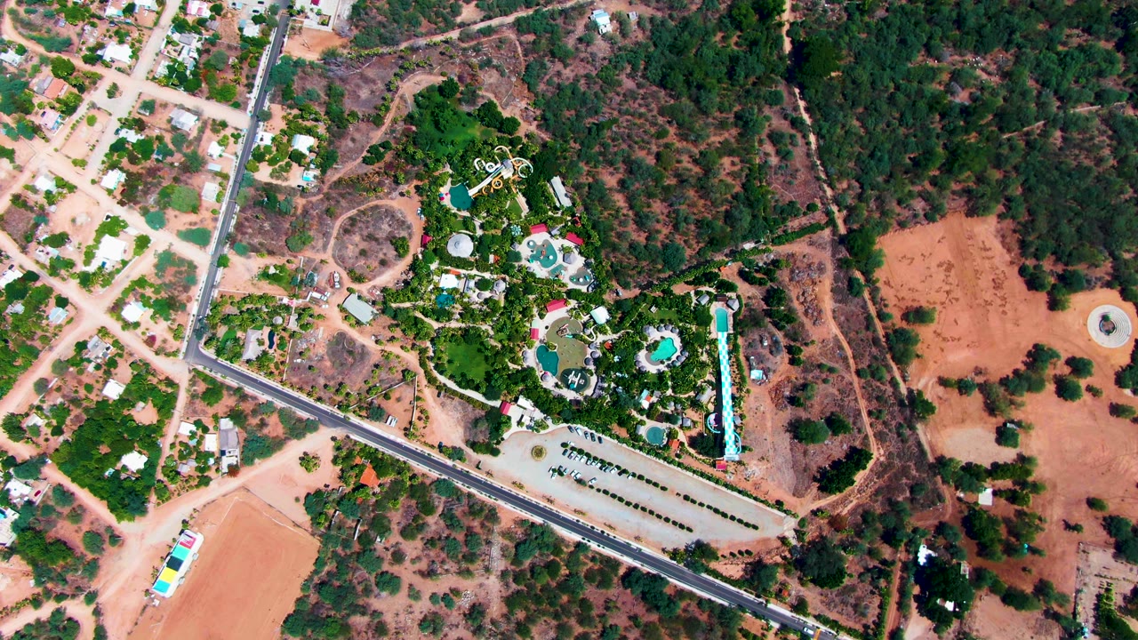 Top-down aerial frame in Caduano with turquoise pools centered among trees, a diagonal paved road, a parking area, dry open ground, and no visible people.