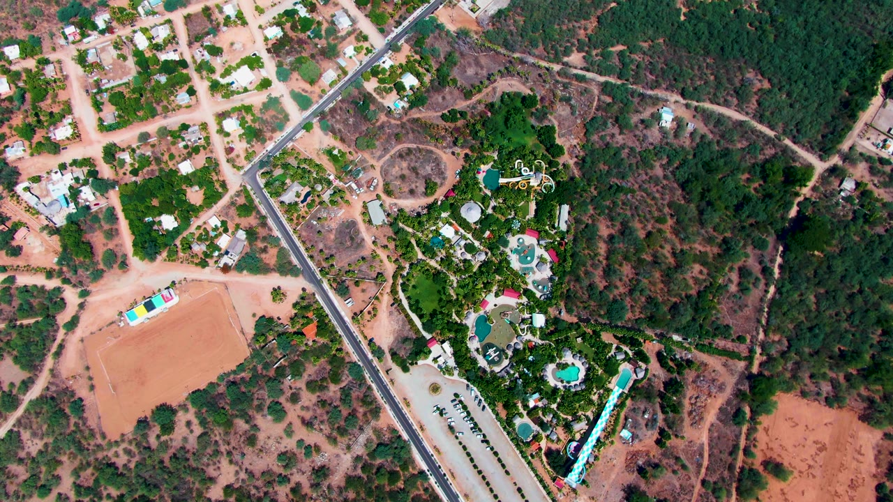 Overhead view in Caduano, Baja California Sur, Mexico showing a diagonal paved road, scattered houses, green pool grounds, reddish dirt fields, and no visible people at the start of the clip.