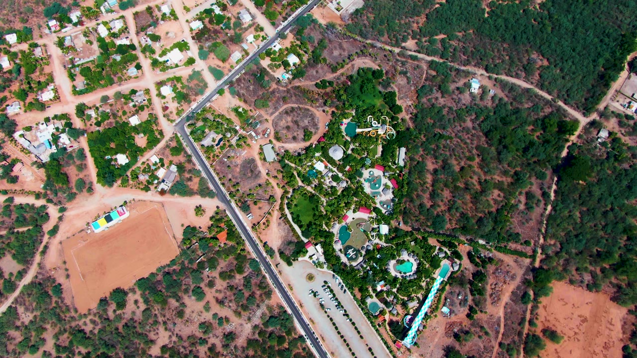 Aerial frame in Caduano with the paved road running diagonally between residential lots, landscaped pool greenery, dry fields, and no visible people one second into the clip.
