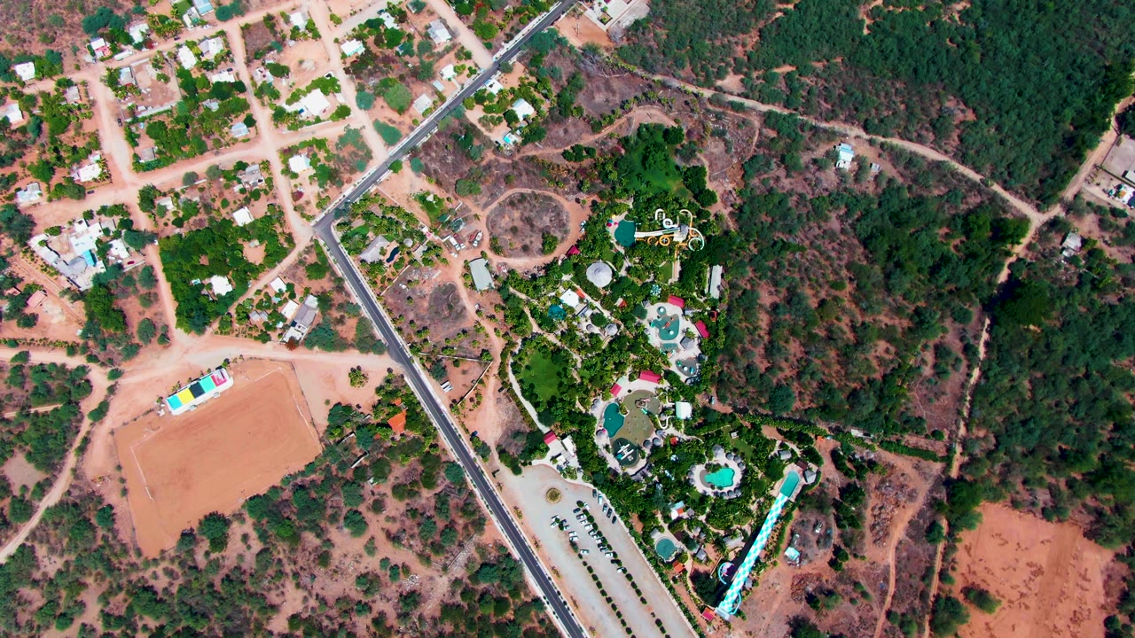 Overhead Caduano frame showing the road beside houses, a compact green pool complex, branching dirt paths, reddish open ground, and no visible people.