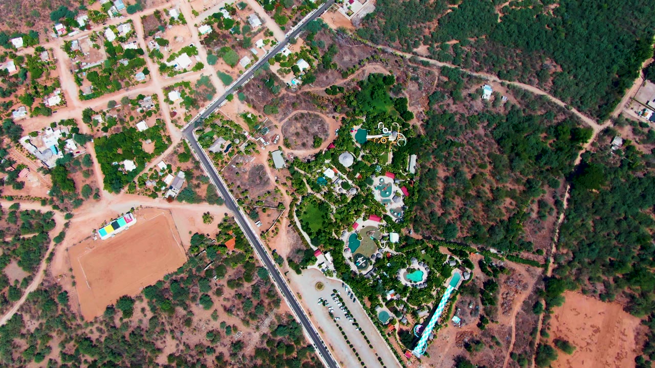 Drone view of diagonal road, housing, trees, pools, and dirt lots.