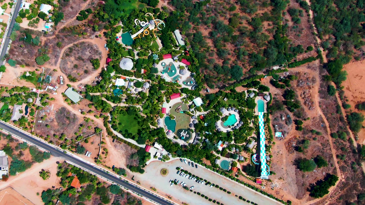 Overhead view of a resort complex in Caduano, Baja California Sur, with turquoise pools, water slides, a road, parking area, dry hills, and no visible people.
