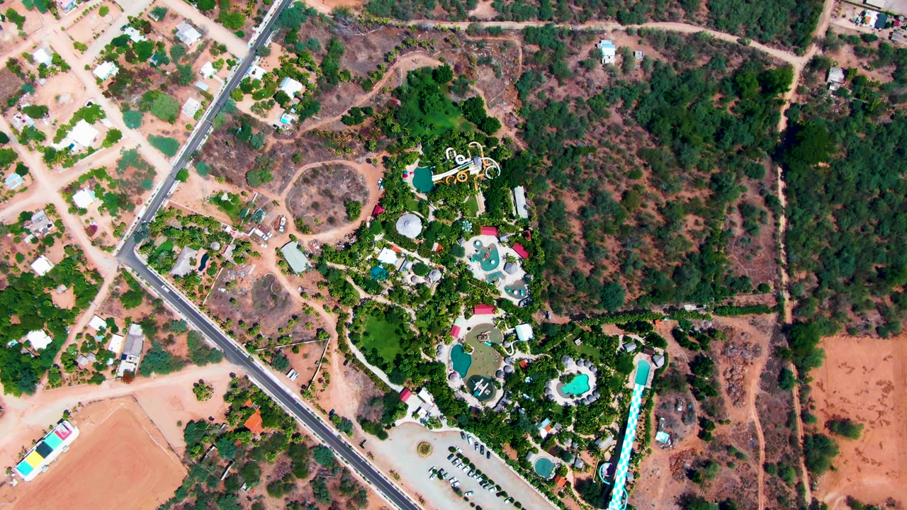 Top-down aerial frame over Caduano, Baja California Sur showing a diagonal paved road, dry parcels, green trees, scattered houses, and blue pools with no visible people.