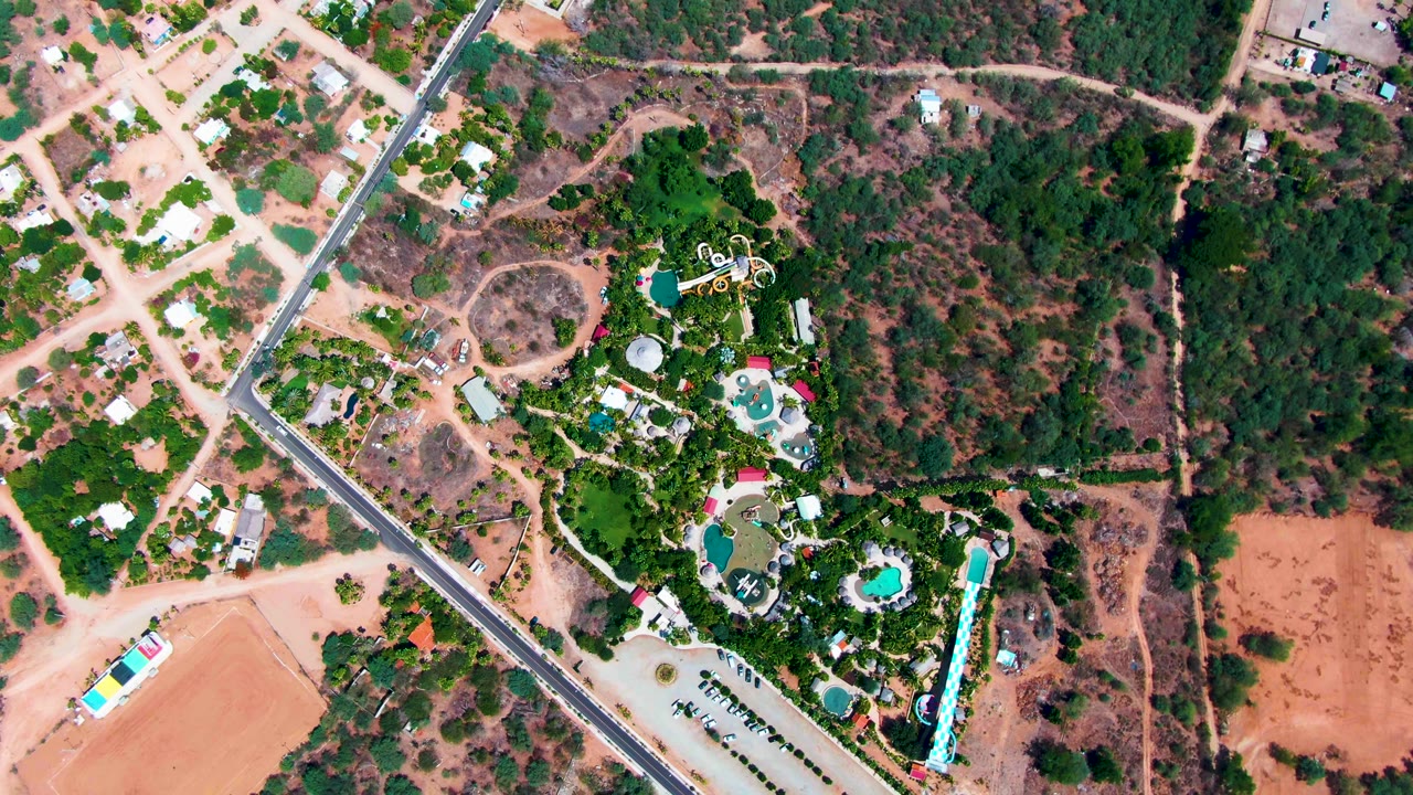 Aerial view over Caduano with the paved road crossing the left side, open dirt lots below, dense trees to the right, and a pool complex near the lower right with no visible people.