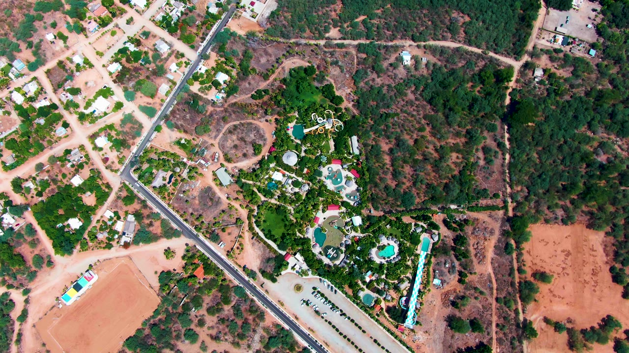 Overhead Caduano frame showing the road running diagonally near houses and scrubland, with landscaped greenery and blue pool shapes on the right and no visible people.