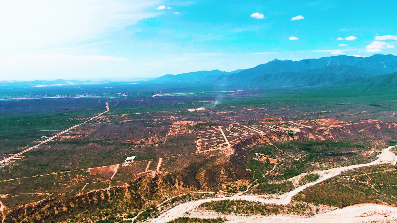 Opening aerial frame over Caduano, Baja California Sur, with pale dirt roads crossing green and brown terrain, a dry wash in the foreground, mountains in the distance, and no visible people.
