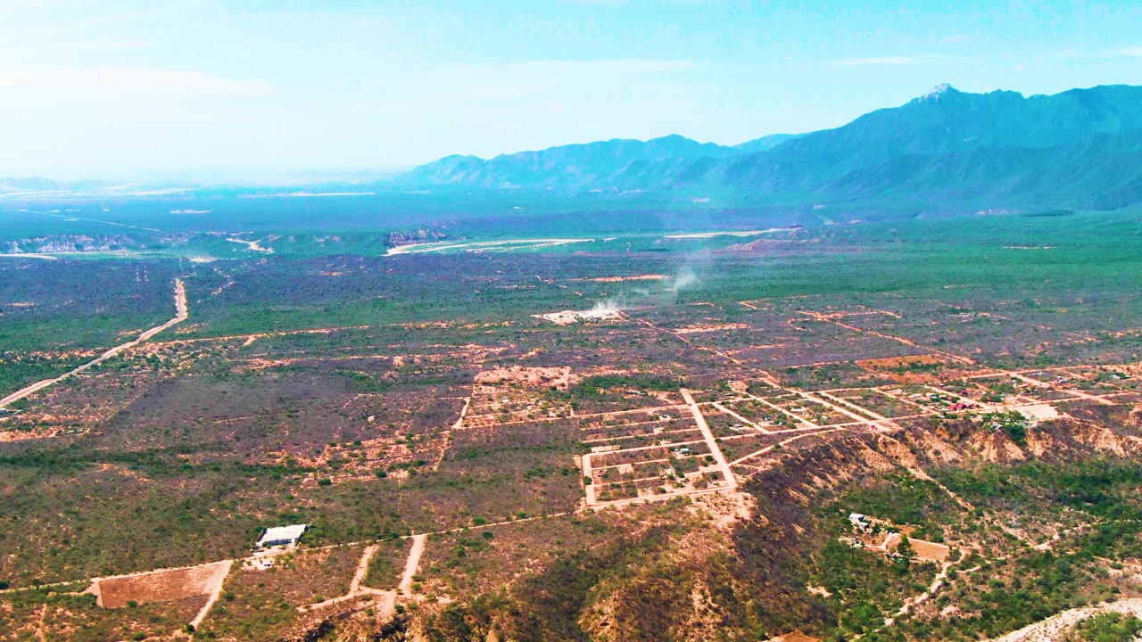 Wide aerial frame over Caduano, Baja California Sur showing a pale smoke plume above mixed green and brown terrain, dirt roads, distant mountains, blue sky, and no visible people.