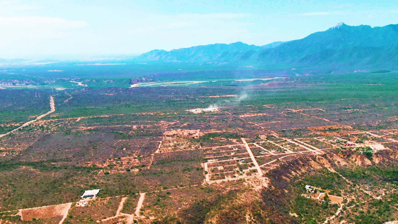 Second aerial frame in Caduano, Baja California Sur with the smoke plume still visible over scrubland, pale roads crossing the midground, mountains on the horizon, and no visible people.