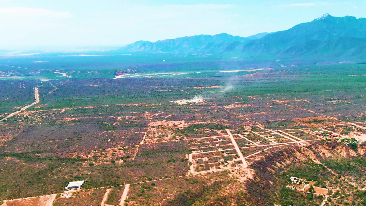 Aerial view over Caduano, Baja California Sur with smoke thinning above dry rural land, a bright road line through the midground, distant mountains, clear sky, and no visible people.