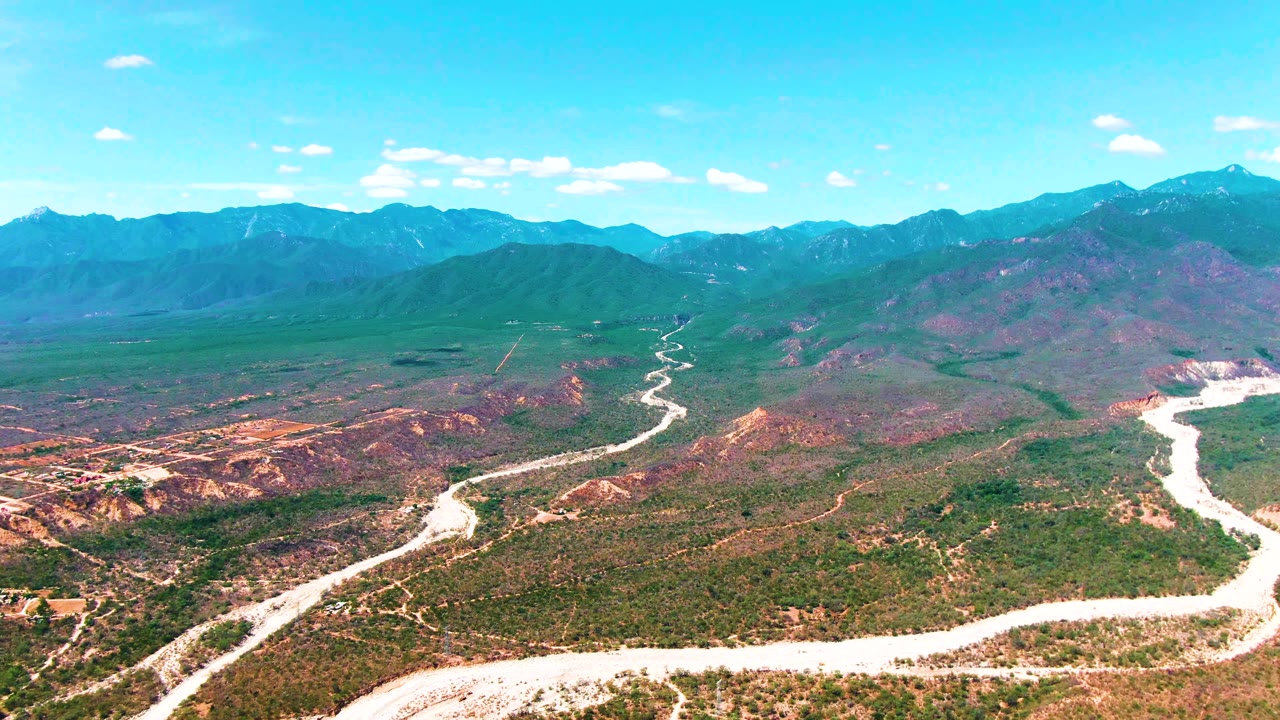 Aerial view over Caduano, Baja California Sur, with a pale winding road crossing green and brown valley terrain toward distant mountains, no visible people.