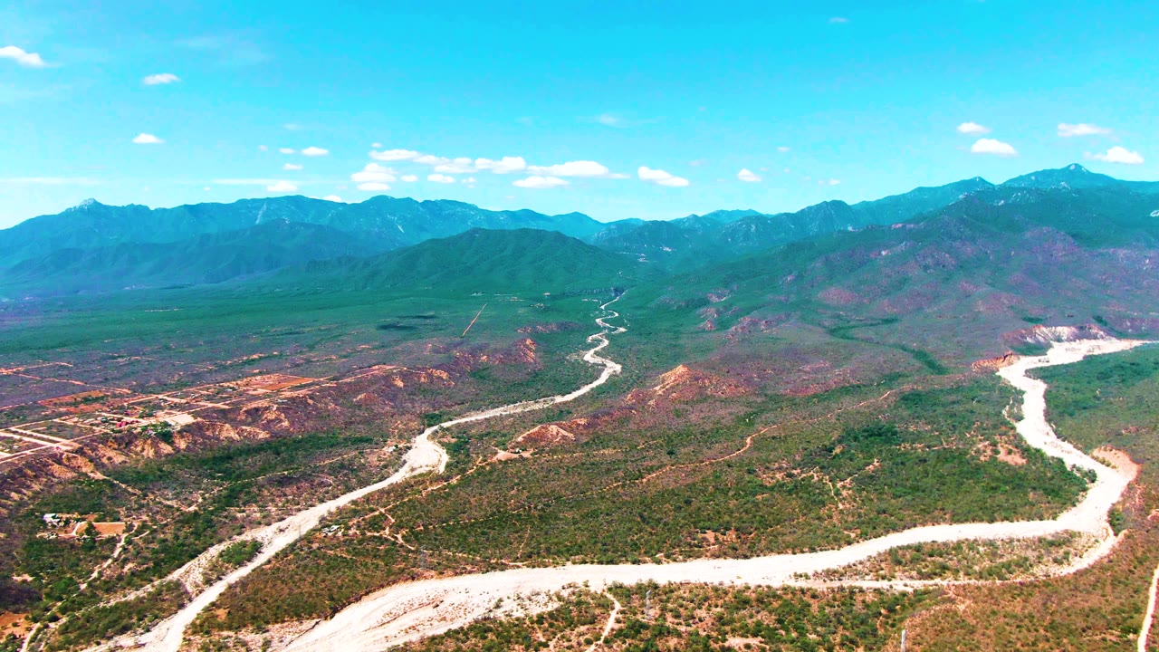 High aerial landscape over Caduano showing the winding pale road across the lower frame, dense green brush patches, brown ridges, and a mountain horizon, no visible people.