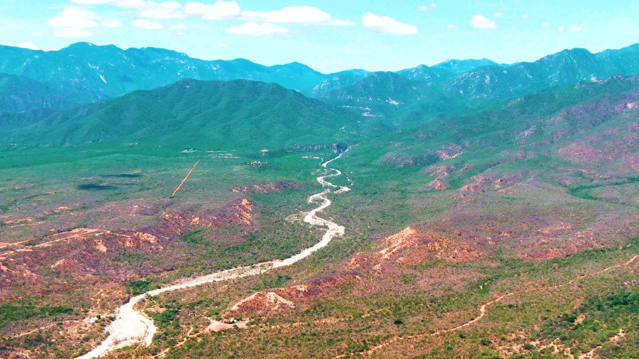 Aerial frame over Caduano valley terrain with the winding road centered lower in the image and green hills rising into blue mountain ridges, with no visible people.
