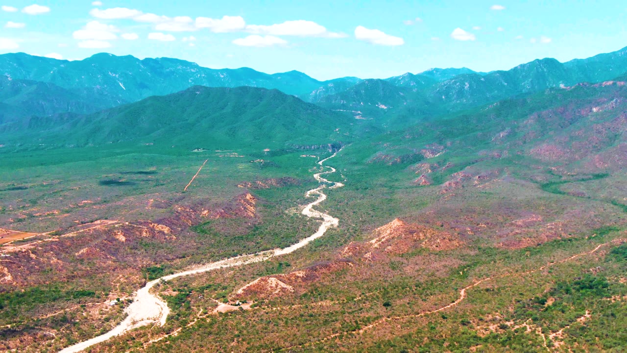 Wide aerial view in Caduano, Baja California Sur, with a bright road bend in the foreground and layered green and blue mountains across the background, with no visible people.