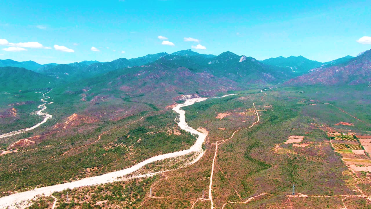 A wide Caduano aerial view shows the river bending through low green-brown terrain, a dirt road running near the channel, distant mountains, scattered clouds, and no visible people.