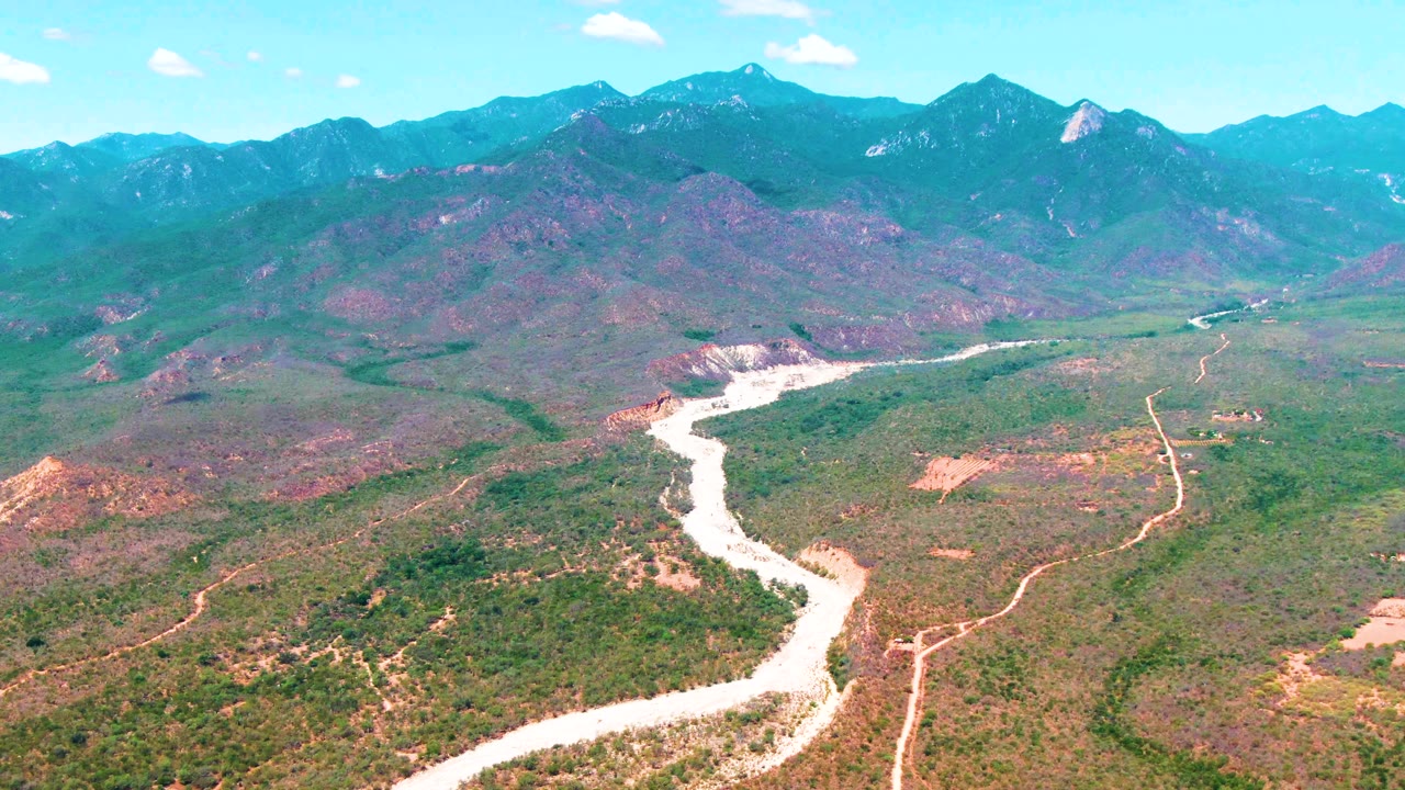Aerial frame over Caduano, Baja California Sur, showing a pale river winding through green and brown valley terrain toward distant mountains, with no visible people.