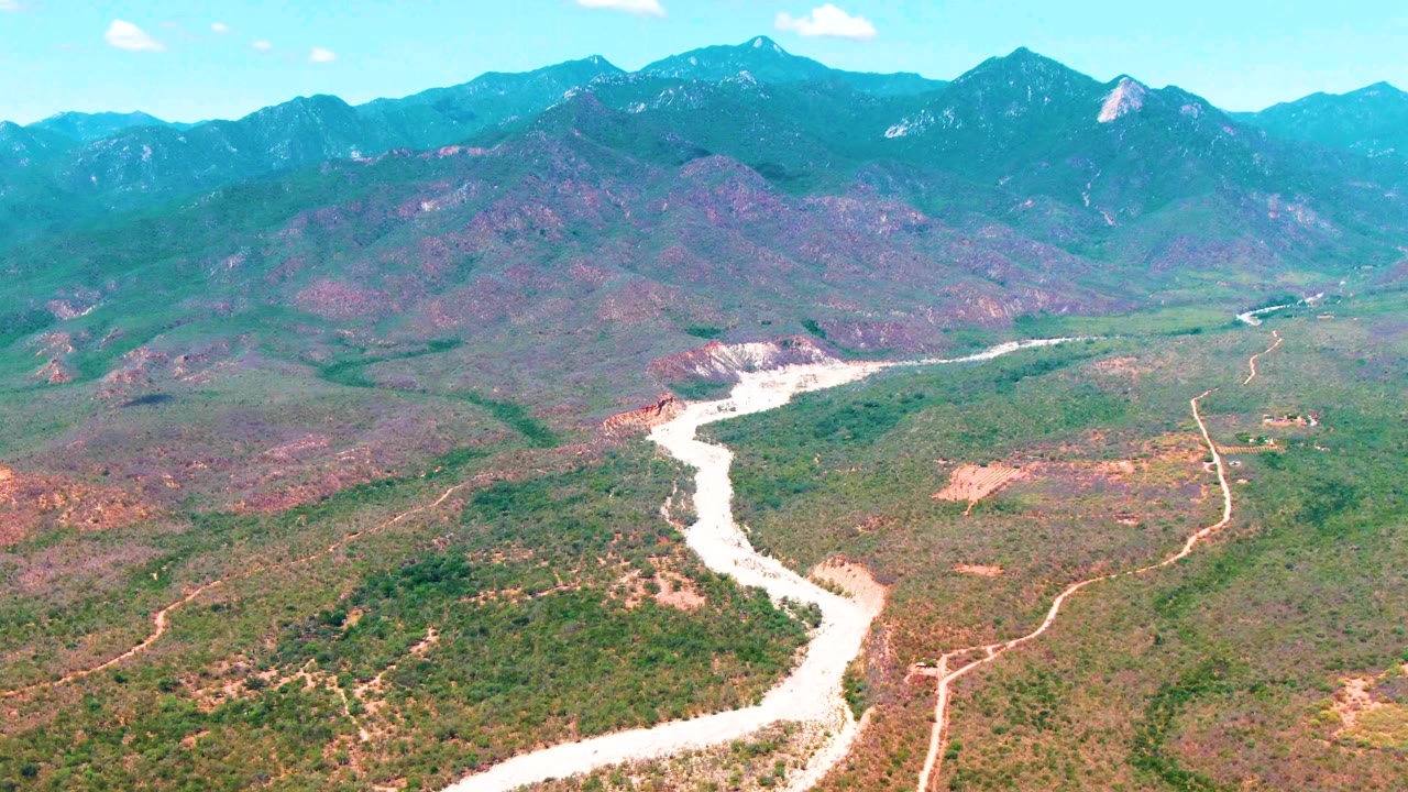 Wide aerial view in Caduano, Baja California Sur, with the riverbed curving from the lower frame into the valley and a broad mountain range under blue sky, with no visible people.