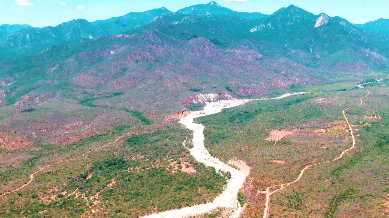 Aerial landscape in Caduano, Baja California Sur, showing a pale river channel crossing the valley beside dirt roads and scrub-covered hills, with no visible people.