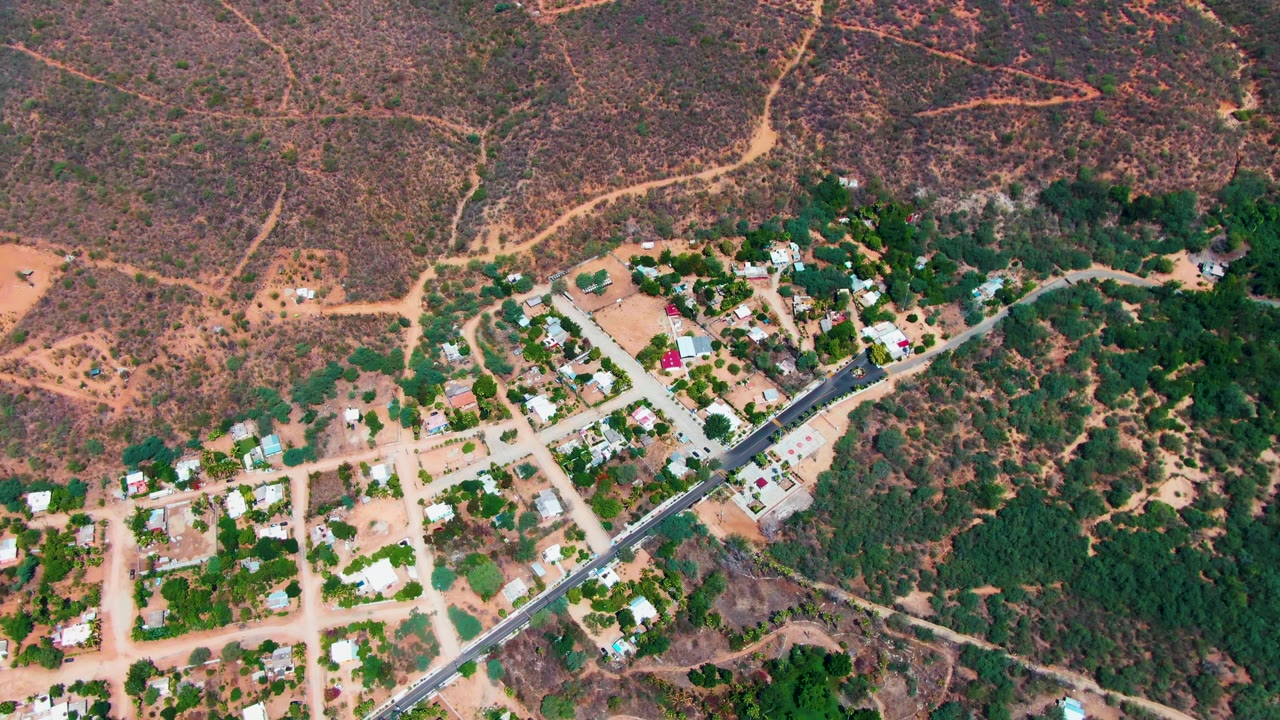 Aerial frame over Caduano, Baja California Sur, showing a diagonal paved road, small homes, dirt side streets, green trees, dry hillside terrain, and no visible people.