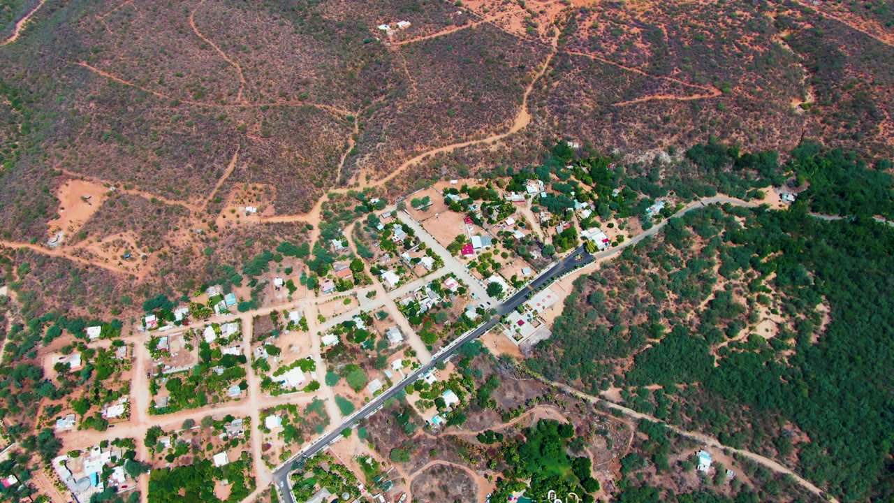 Caduano aerial frame showing a loose grid of dirt streets, scattered homes, a diagonal paved road, dense green vegetation on the right side, dry reddish hills beyond, and no visible people.