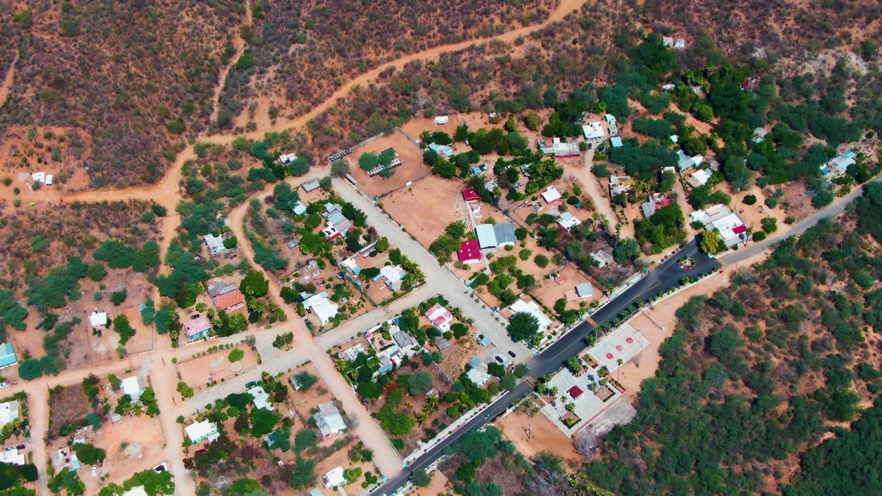 Aerial frame over Caduano, Baja California Sur, showing scattered homes, a paved road, dirt paths, green tree clusters, and dry hillside brush, with no visible people.