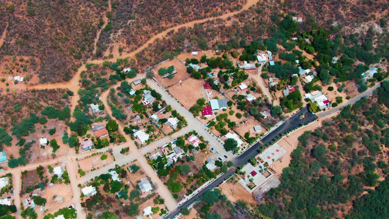Second-frame aerial view in Caduano showing the paved road cutting past small homes, pale lots, trees, and dry hillside tracks, with no visible people.
