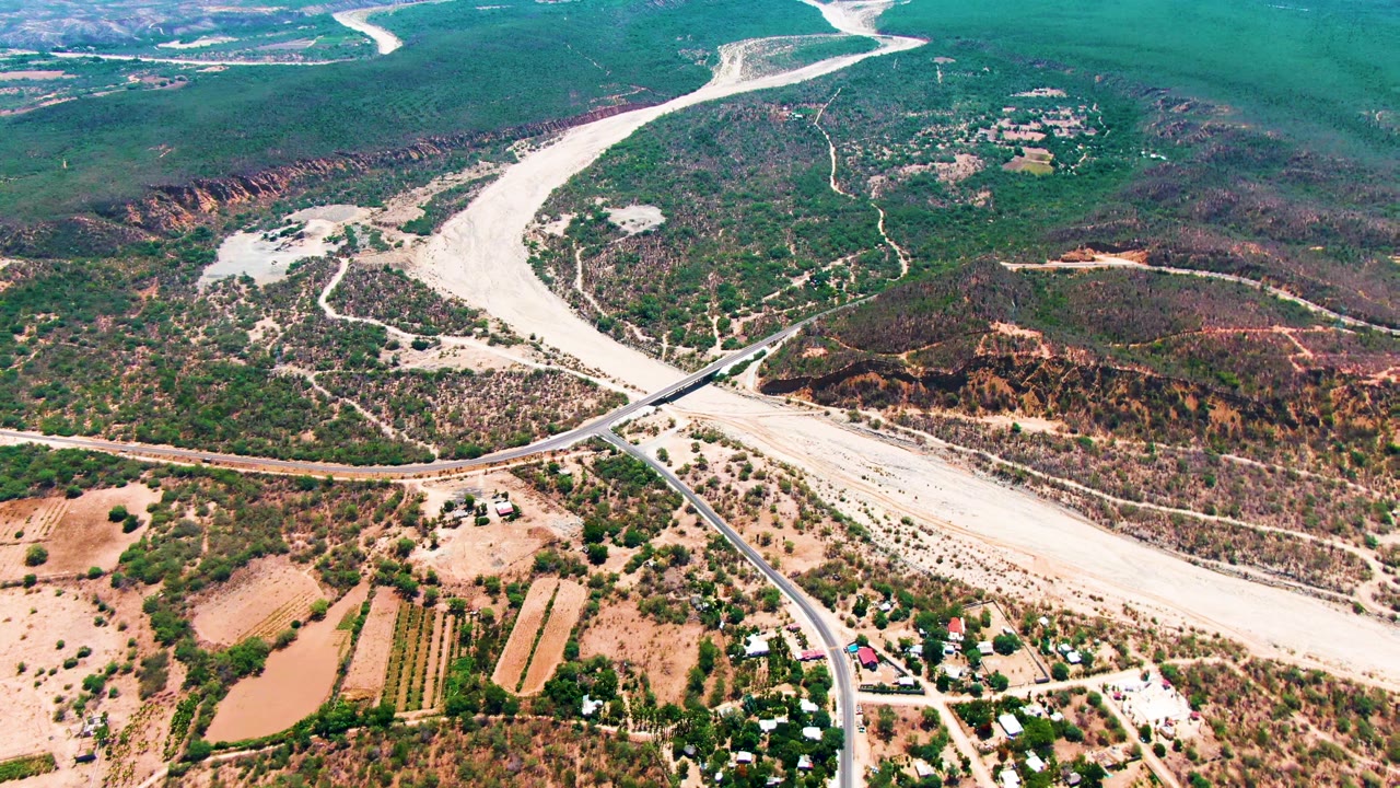 Aerial frame over Caduano, Baja California Sur, with a pale dry riverbed, paved road crossing, scrub hills, fields, scattered buildings, and no visible people.