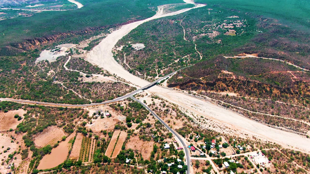 Drone frame over Caduano with the riverbed curving through scrubland, a paved road and vehicle in the midground, cultivated plots below, and no visible people.