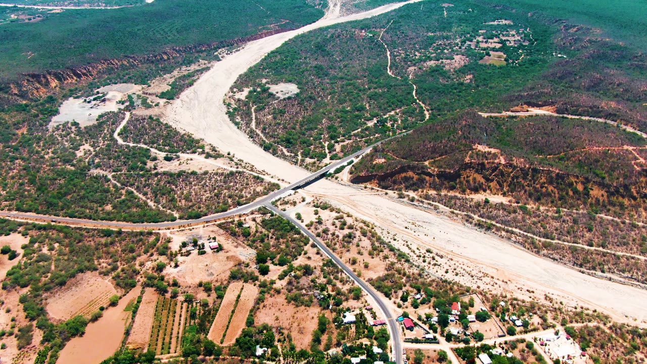 Paved road crossing a pale riverbed below green and brown hills.