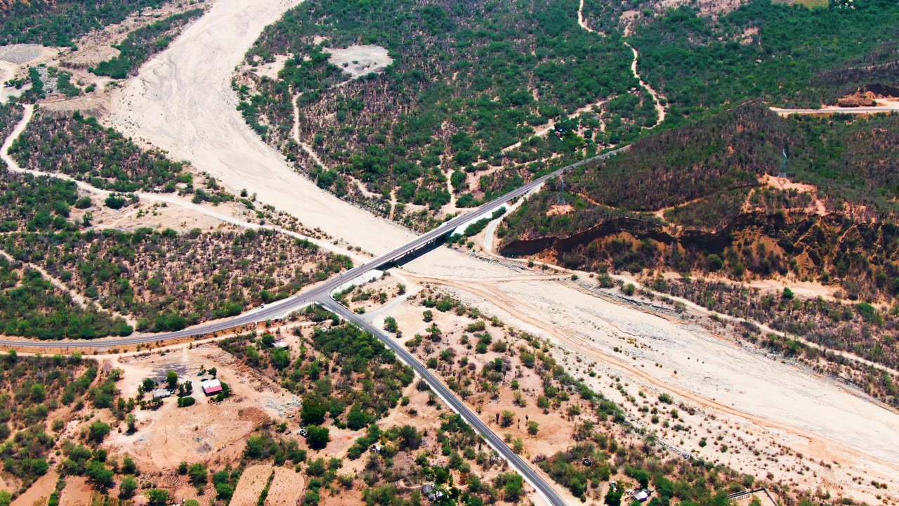 Aerial frame from Caduano, Baja California Sur, showing a paved crossroad and bridge over a pale dry riverbed with sparse green vegetation and no visible people.
