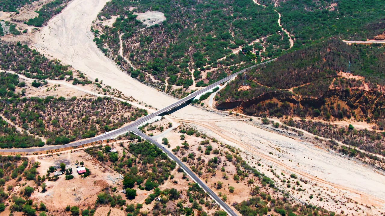 Aerial frame from Caduano, Baja California Sur, with vehicles on the paved road beside a broad dry wash, scrub vegetation, reddish exposed soil, and no visible people.