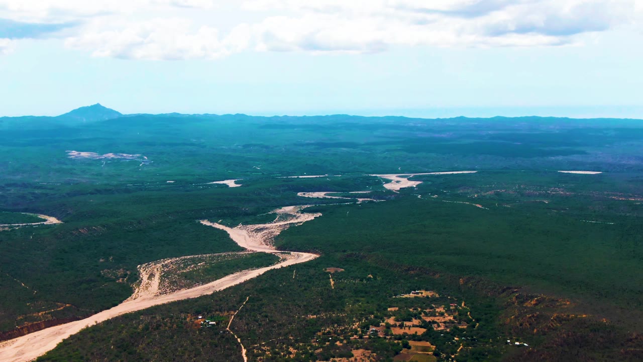 Wide aerial view of a sandy river winding through forest toward distant mountains.
