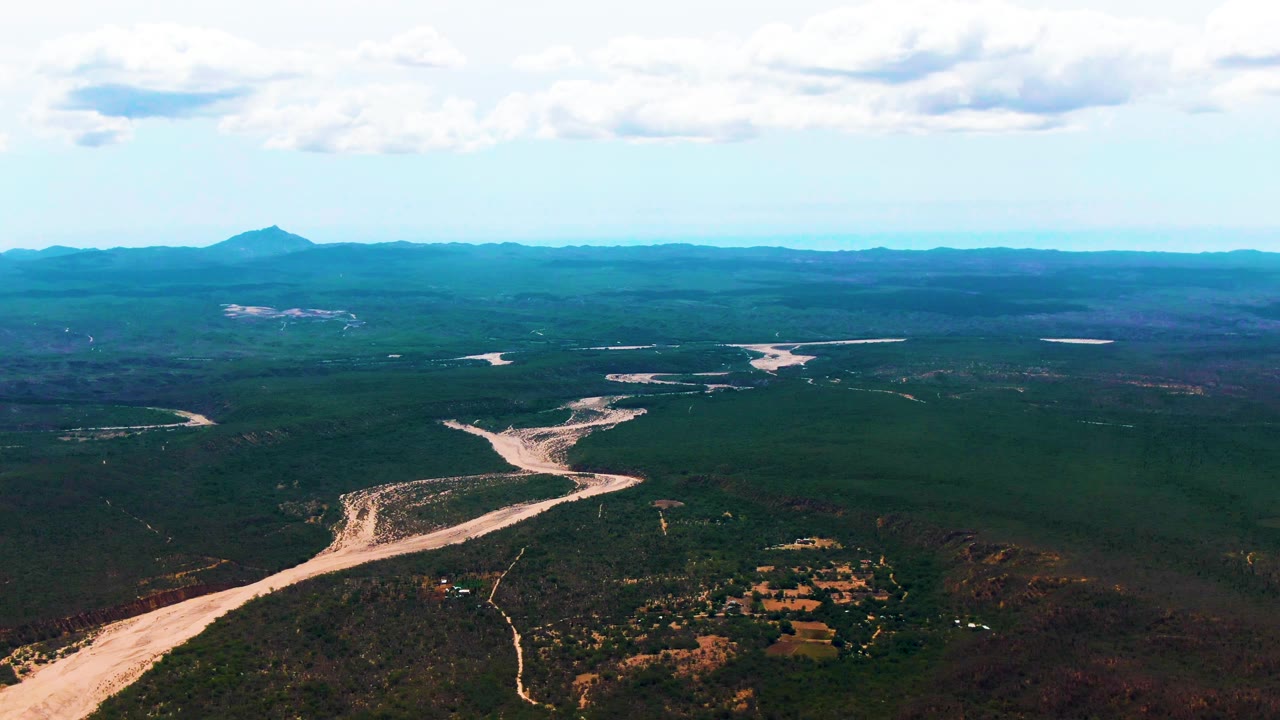 At 0:03, the wide Caduano drone view shows the sandy riverbed entering from the lower left, crossing forested terrain, and receding toward a mountain horizon, with no visible people.