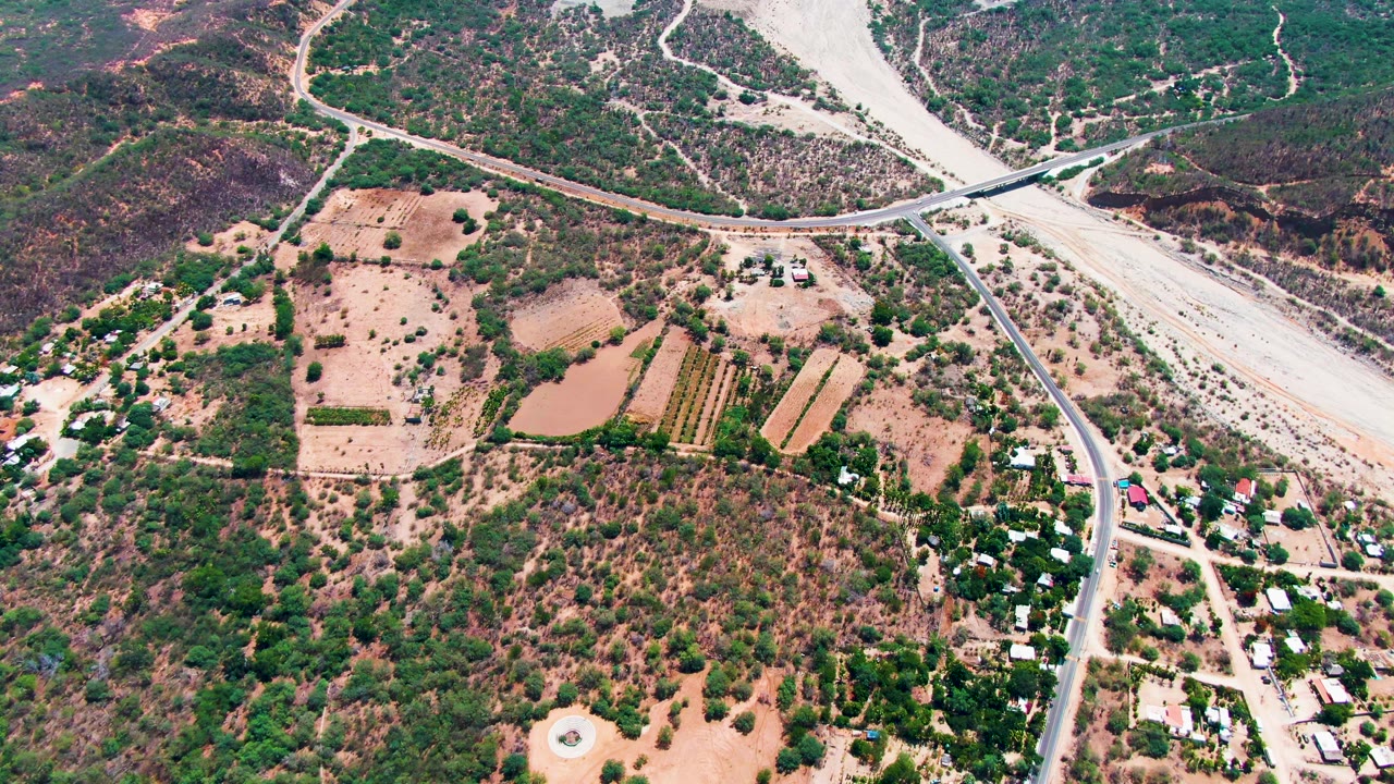Aerial frame over Caduano, Baja California Sur with a rural road crossing near a dry riverbed, tan fields, green scrub, scattered small buildings, and no visible people.