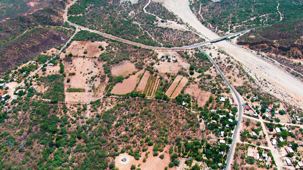 High aerial view in Caduano, Baja California Sur showing tan agricultural plots, a small brown pond, scrub-covered hills, a paved road, a dry riverbed, and no visible people.