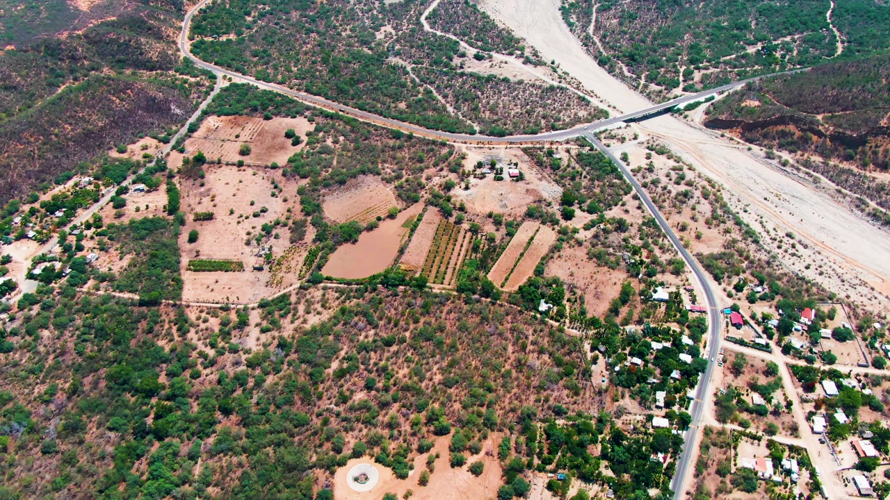 Drone frame over Caduano, Baja California Sur with the road and bridge near the top, dry farmland and a small pond below, scattered houses to the right, and no visible people.