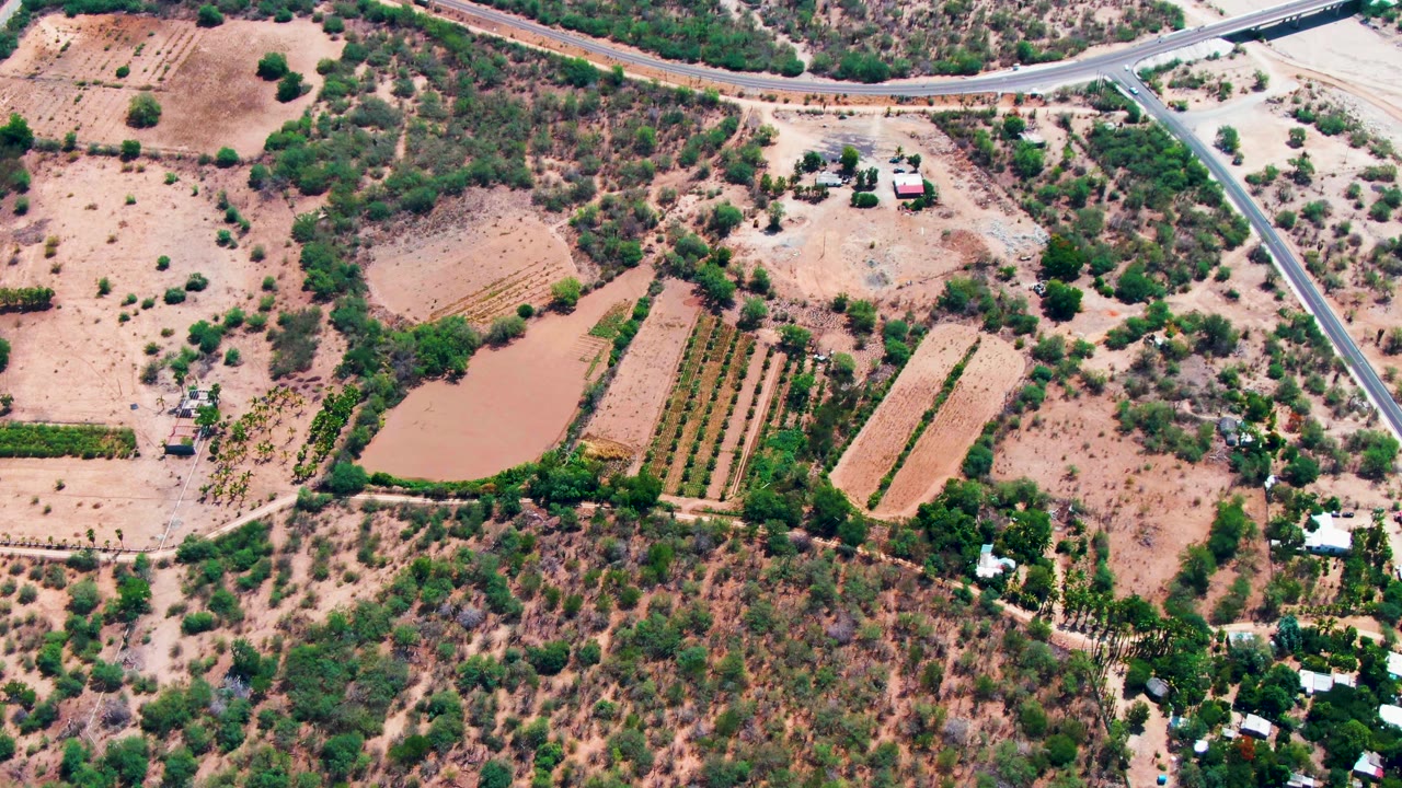 Aerial frame over Caduano farmland with tan tilled plots, green crop rows beside a muddy pond, crossing paved roads, small structures, and no visible people.