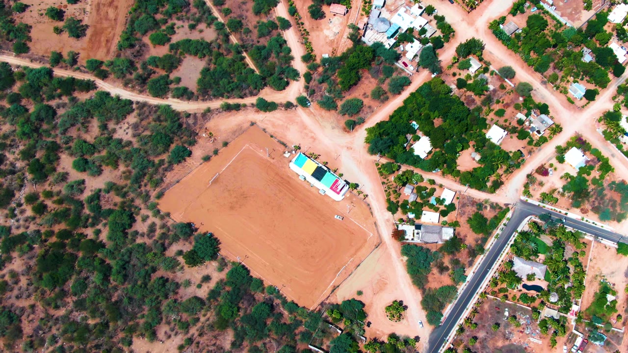 Top-down aerial view of Caduano, Baja California Sur with a tan rectangular dirt field, scattered houses, dirt roads, green trees, and no visible people.
