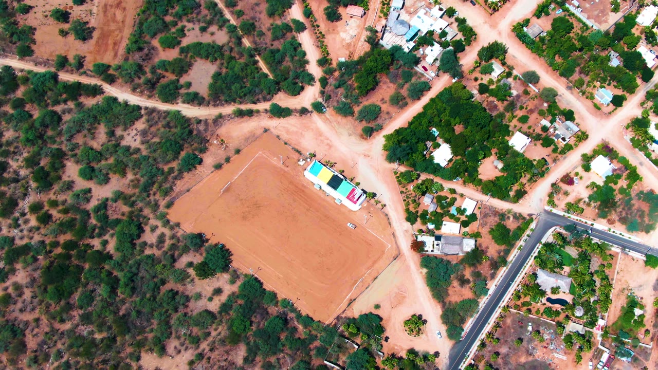 A slightly shifted overhead frame shows Caduano houses, pale dirt tracks, a large dirt field, dense green tree patches, and no visible people.
