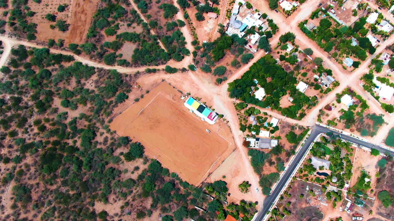 The overhead drone view in Caduano shows a tan field at left, intersecting dirt roads, clusters of small buildings, scrubby vegetation, and no visible people.