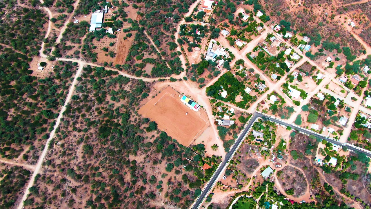 Overhead frame from Caduano, Baja California Sur showing a reddish dirt lot, pale roads, scattered houses, dry brush, and a white vehicle near the start of the clip, with no visible people.