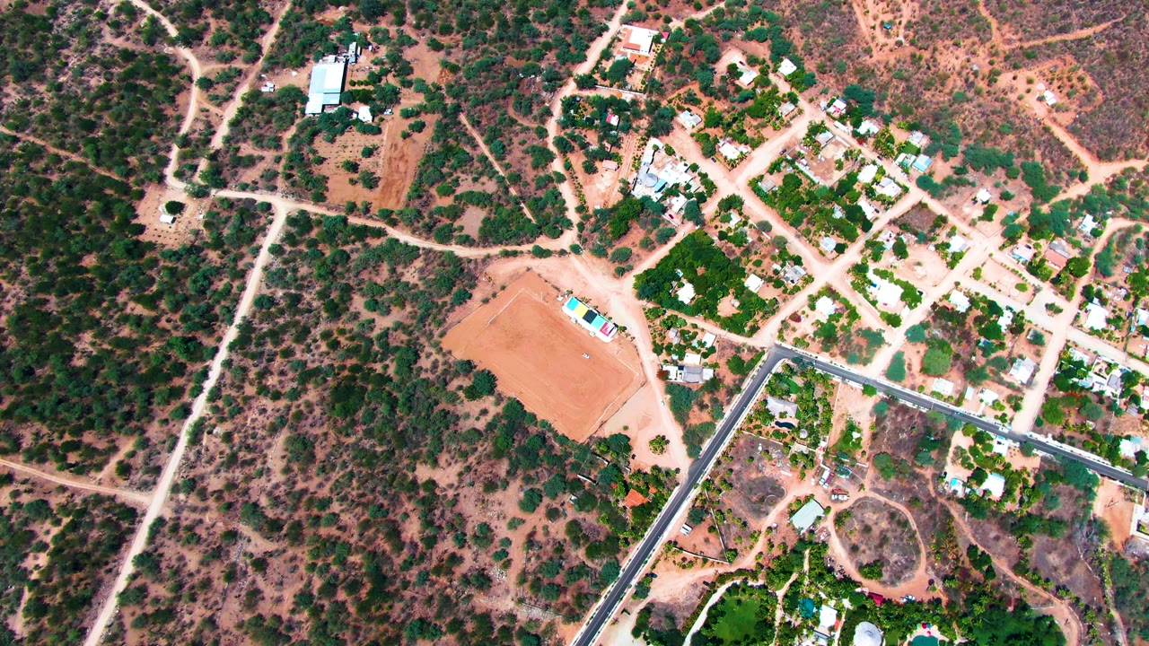Overhead frame from Caduano, Baja California Sur with the empty reddish lot at left, a grid of light dirt roads, small residential structures, dry vegetation, and no visible people.