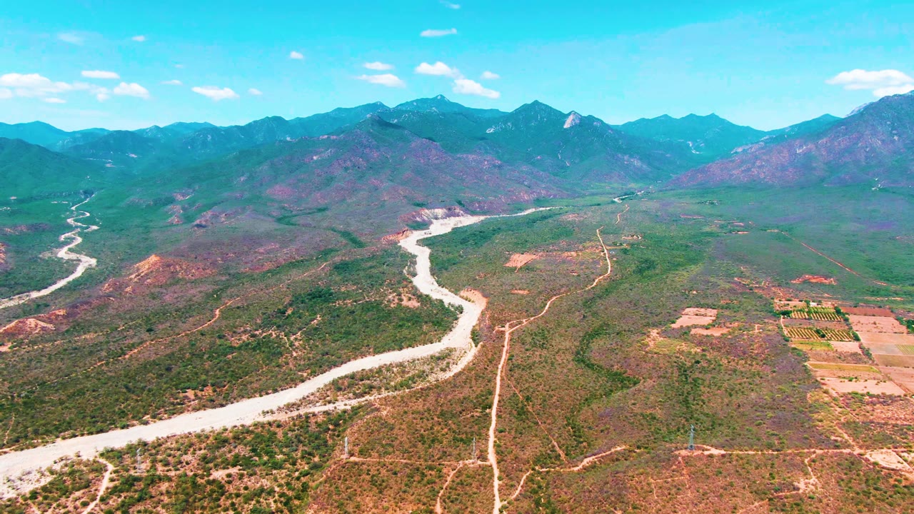 Opening aerial frame over Caduano, Baja California Sur, showing a pale winding river through green and brown valley terrain, cultivated fields at right, distant mountains, and no visible people.