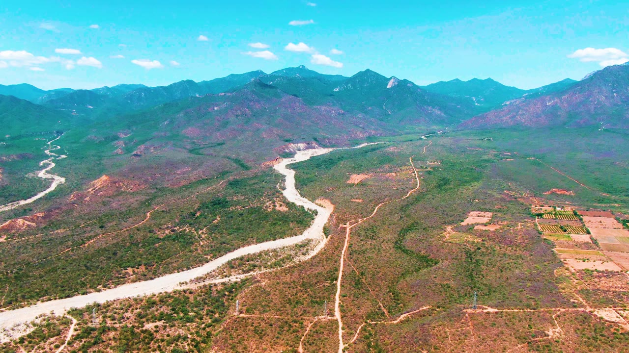 Aerial frame over Caduano, Baja California Sur, with the river bending from the lower left toward the mountain range, field blocks to the right, scattered clouds, and no visible people.
