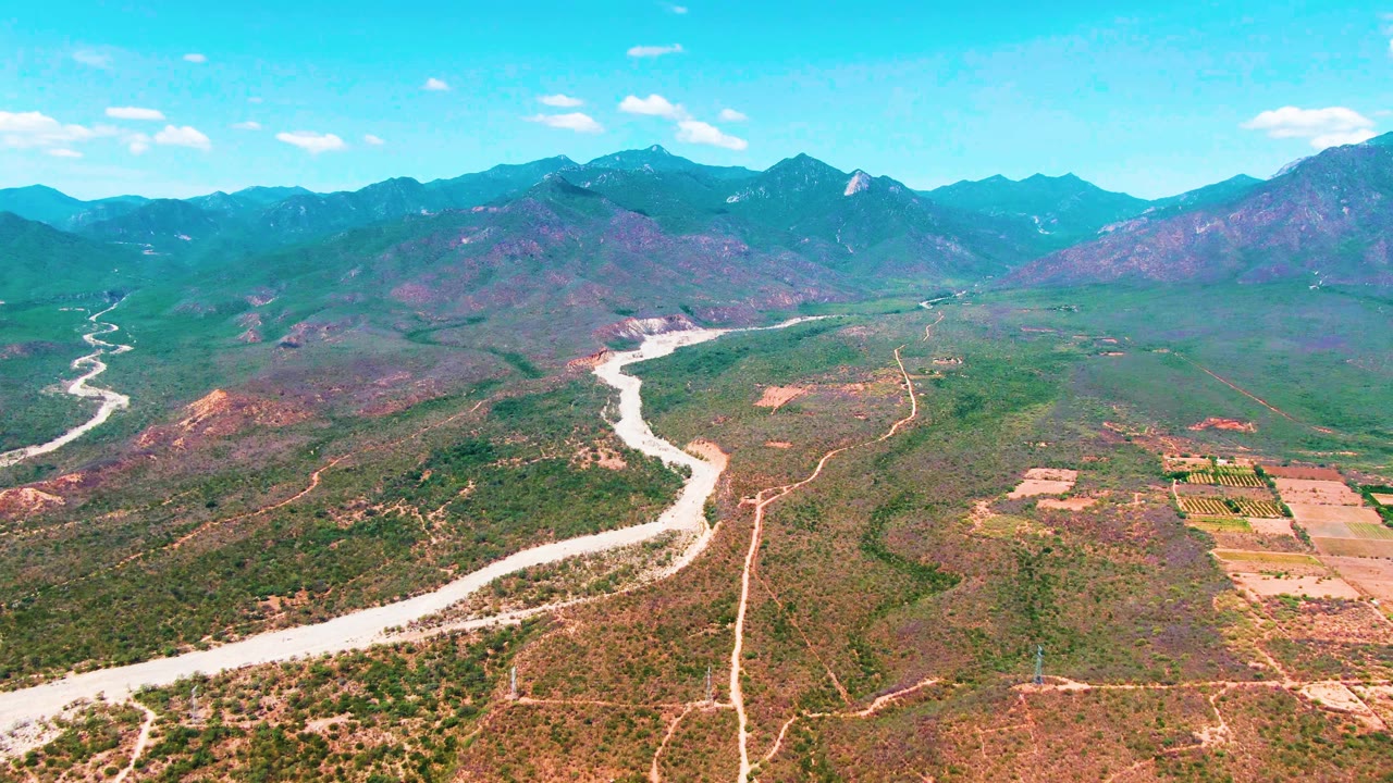 Caduano aerial view showing the bright river channel crossing the valley floor, tan dirt roads, rectangular fields on the right, layered mountains in the distance, and no visible people.