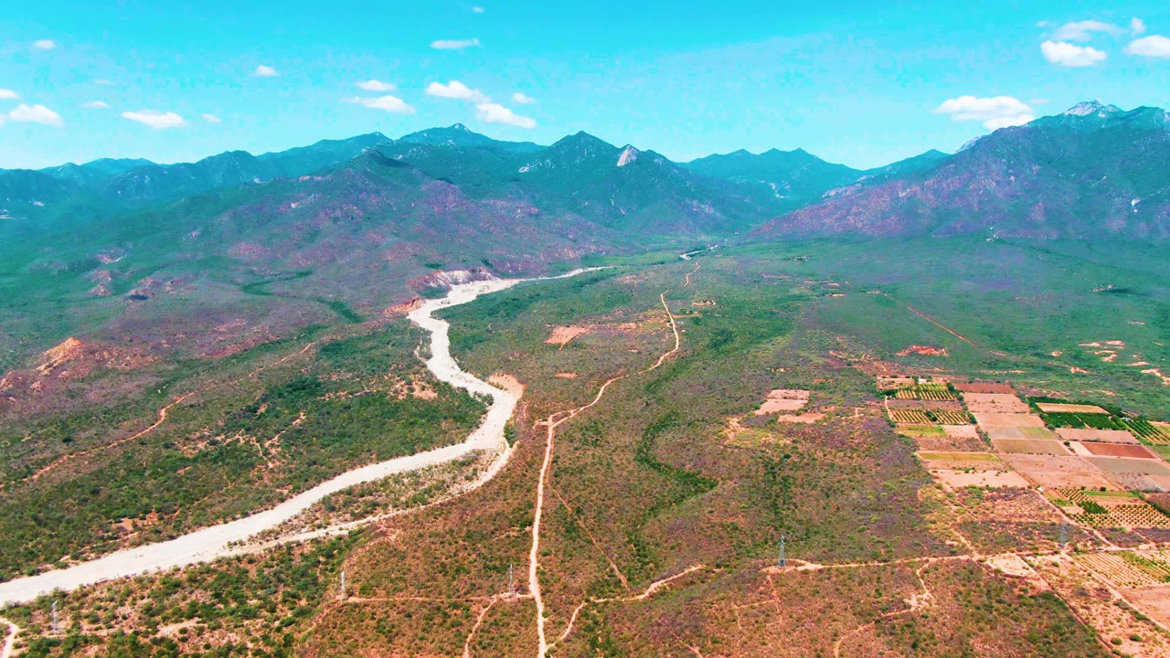 Aerial frame in Caduano, Baja California Sur, showing the river curve slightly farther through the valley, with rectangular fields to the right, mountain ridges on the horizon, and no visible people.