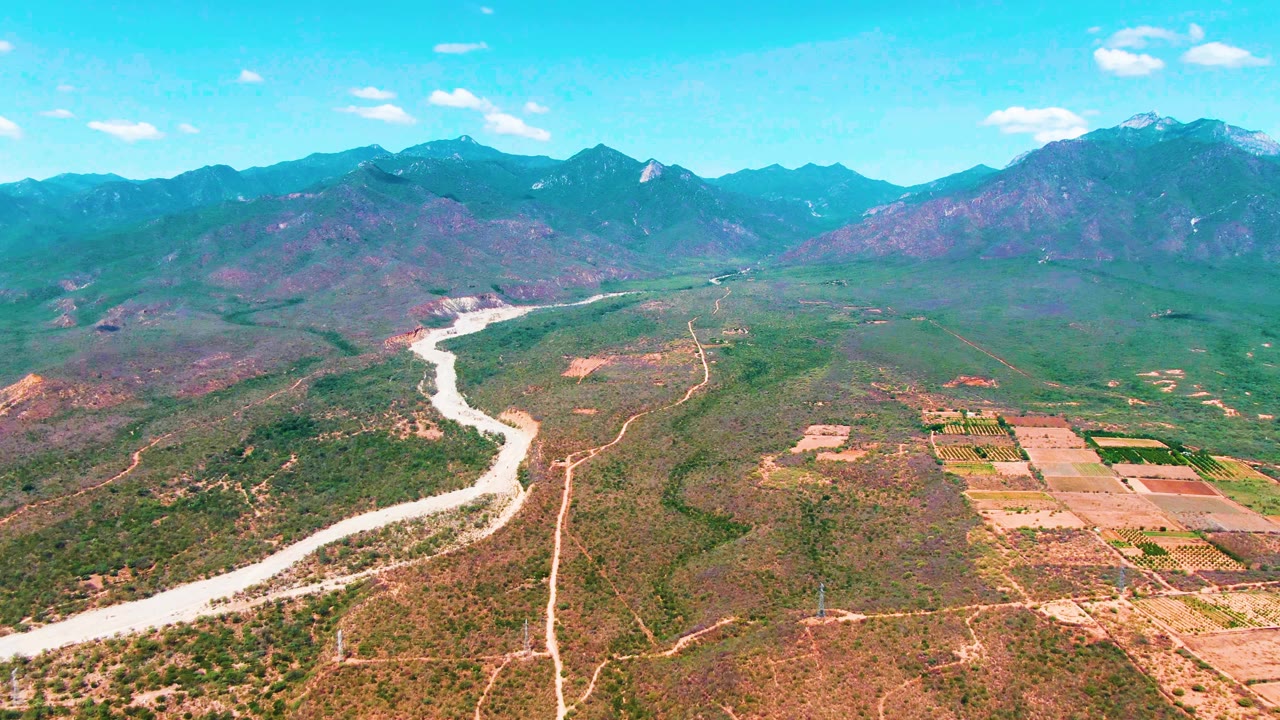 Later frame over Caduano, Baja California Sur, with the river running from the lower left toward the center, fields occupying the right side, broad mountain slopes beyond, and no visible people.