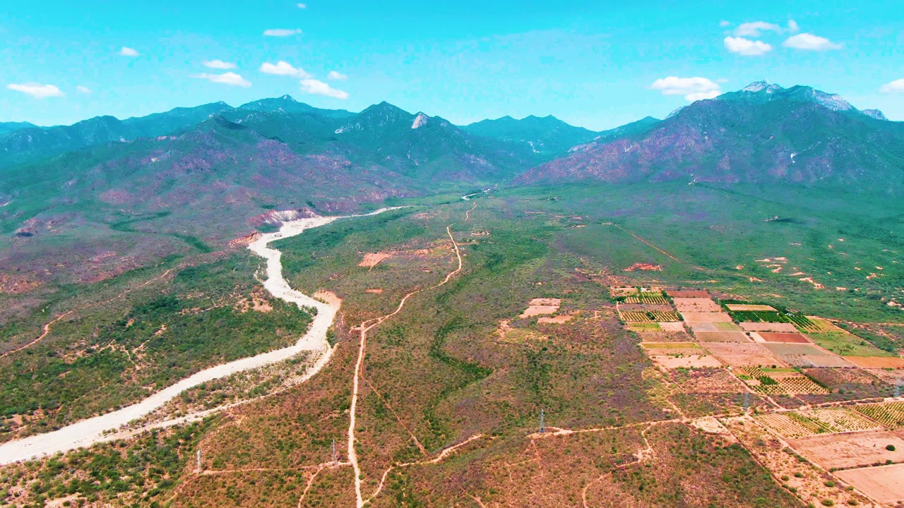 Wide aerial frame over Caduano, Baja California Sur, showing a pale winding river at left, patchwork fields at right, distant mountain ridges, bright blue sky, and no visible people.