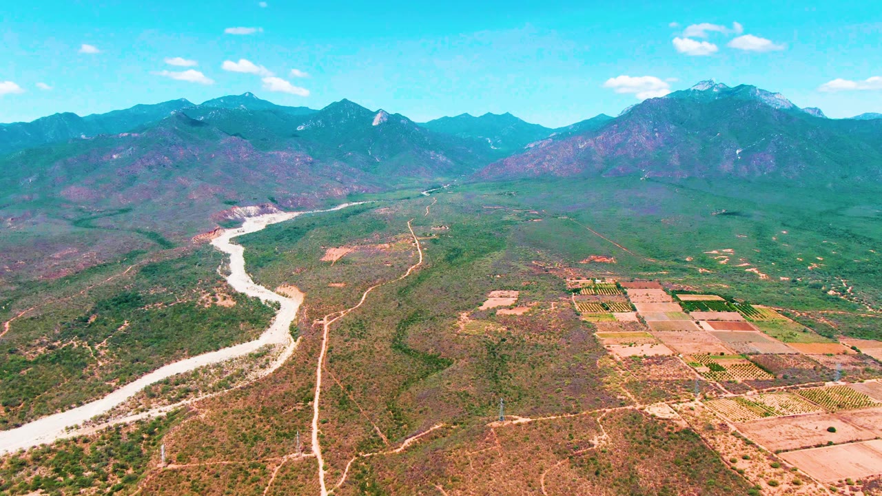 High drone-style view over Caduano, Baja California Sur, where the river curves through the left side, green and brown valley terrain fills the middle, farmland lies to the right, and no visible people appear.