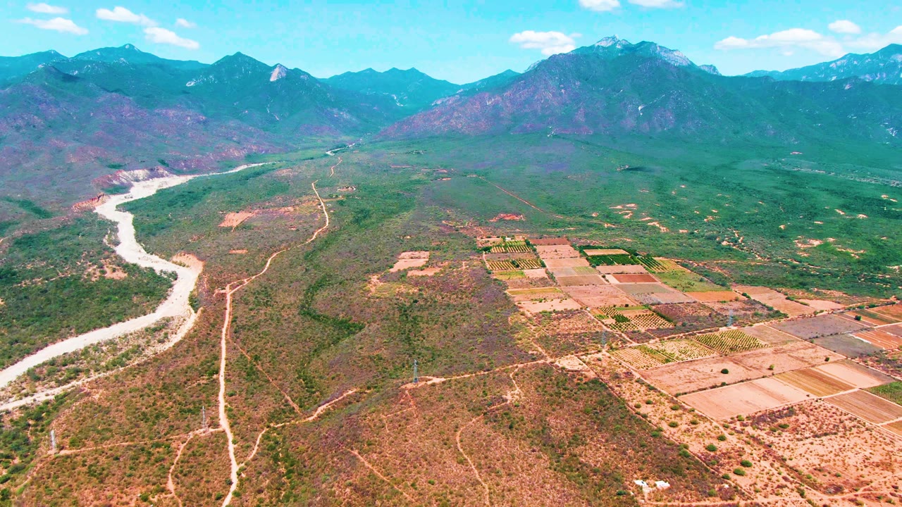 Opening aerial frame over Caduano, Baja California Sur, showing a pale river bending through dry green-brown valley terrain, farm plots to the right, distant mountains, and no visible people.