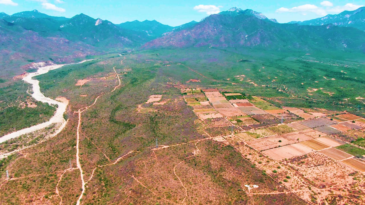 Mid-clip aerial frame over Caduano, Baja California Sur, showing more of the gridded fields at right, pale dirt roads across dry terrain, the river at left, distant mountains, and no visible people.